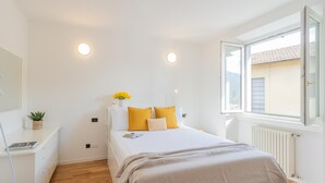 A bedroom with a bed, a bedside table with a plant, a window with a view of buildings, and a radiator. - Ponente Balcony Garden (Gravedona)