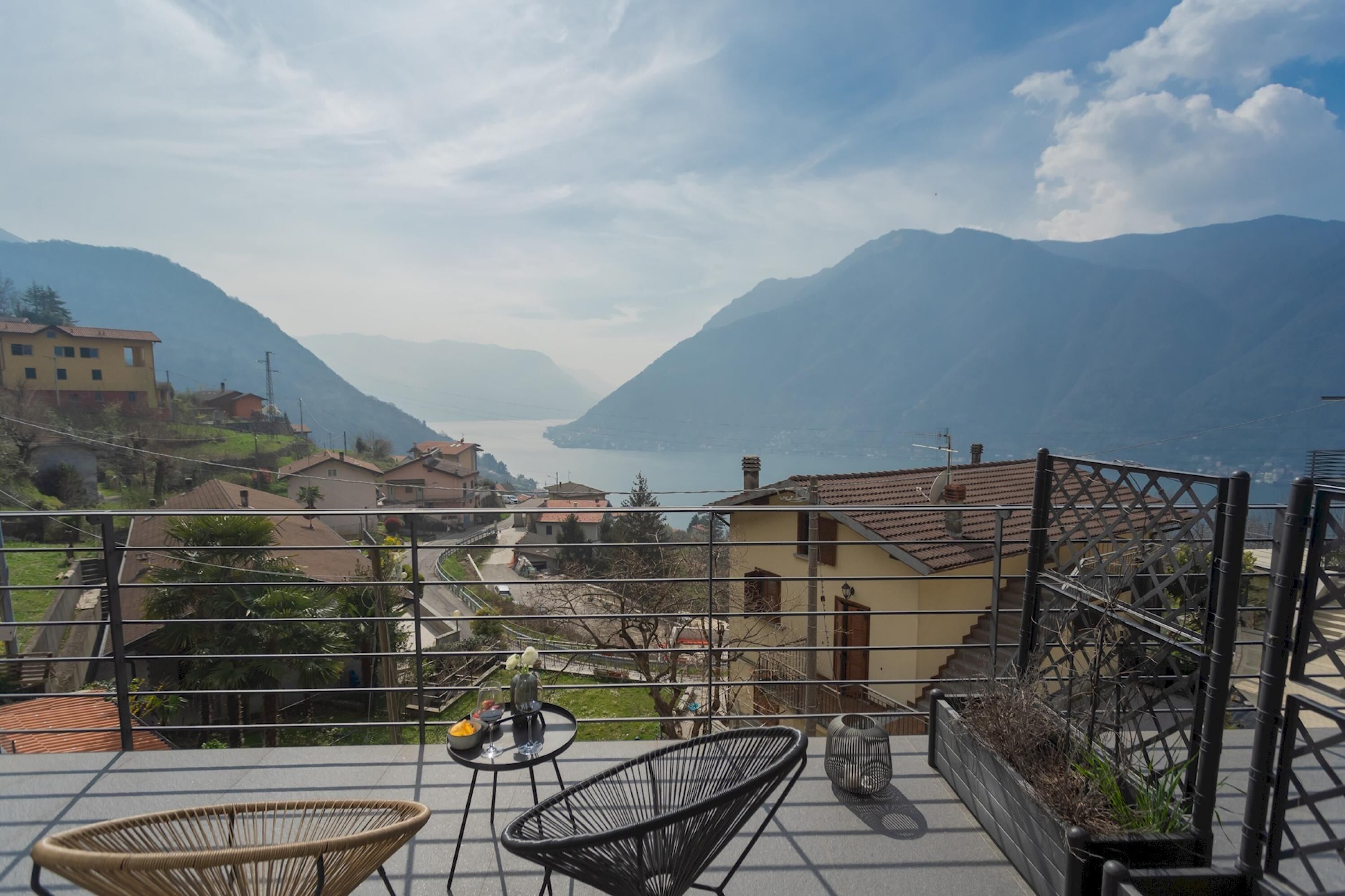 A terrace with wicker chairs, a small table with a vase and wine glasses, and a view of a valley with mountains and a lake.