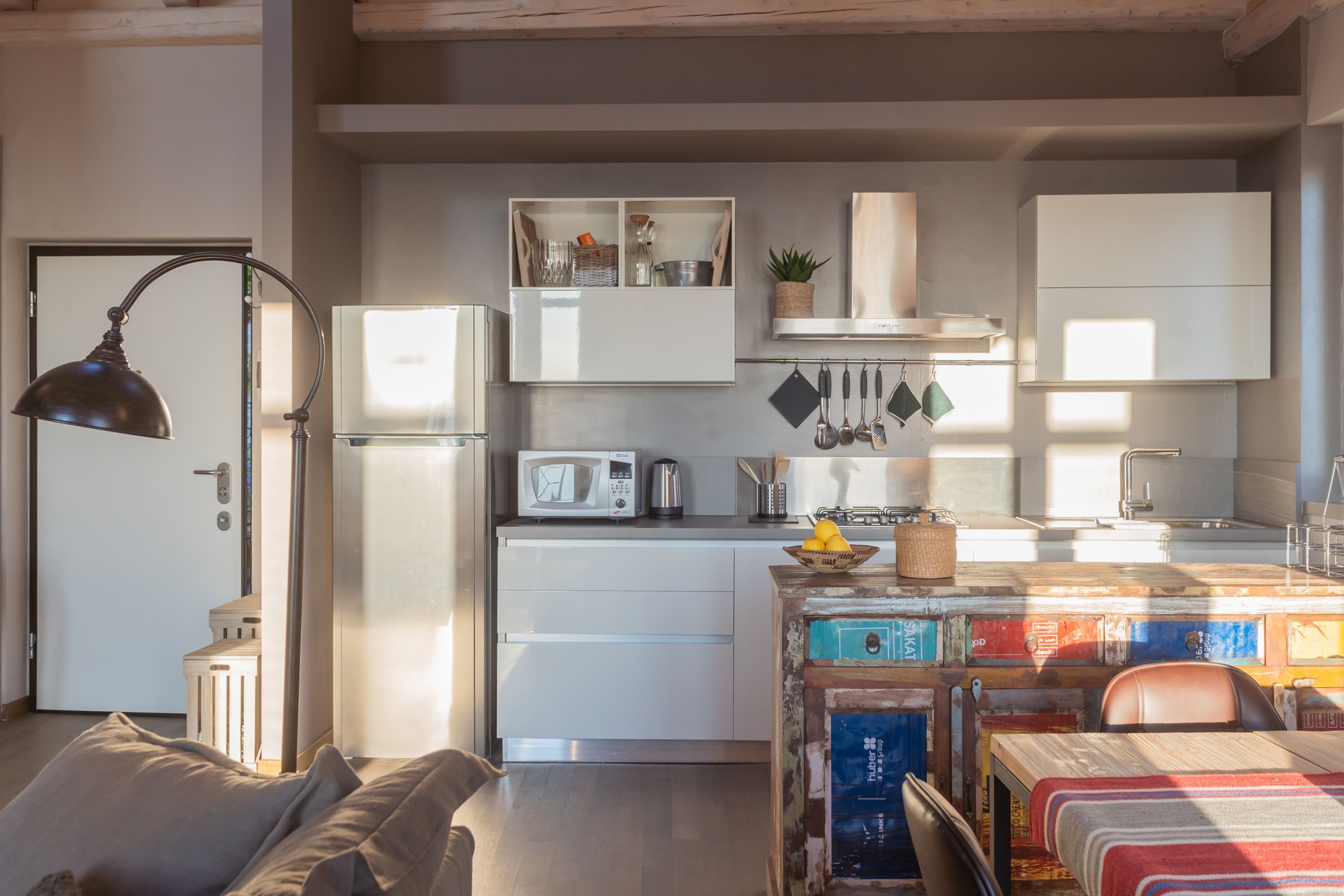 A modern kitchen with white cabinets, a microwave, and a sink. There is a dining area with a table and chairs, and a bed with a lamp.