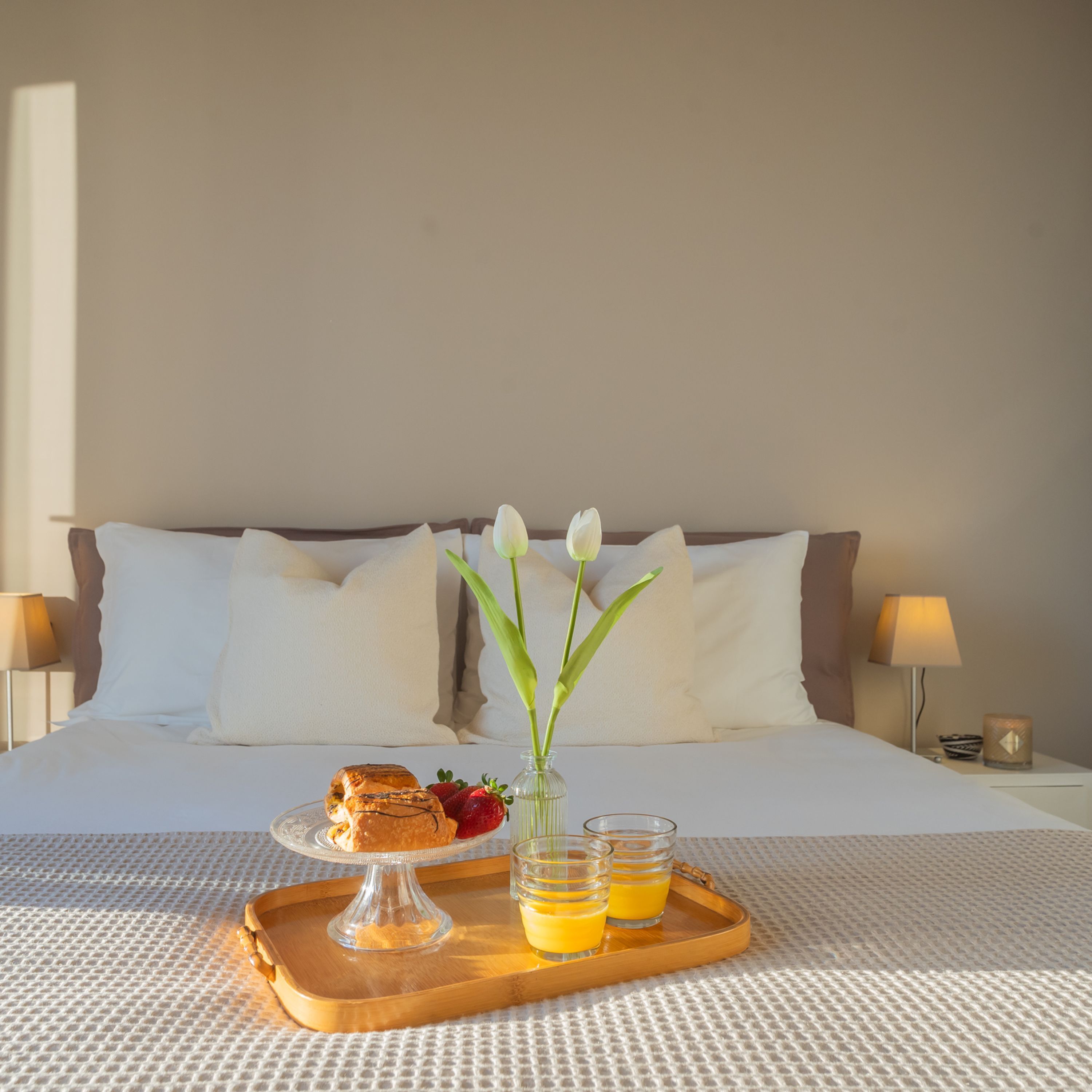 A neatly made bed with white linens, a tray with breakfast items including pastries, strawberries, and orange juice, and a vase with white flowers.