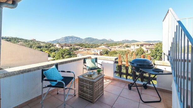 A balcony with a view of a town and mountains, a grill, two chairs, and a small table. - Bilo Porto Rotondo Castello 3 (Olbia)