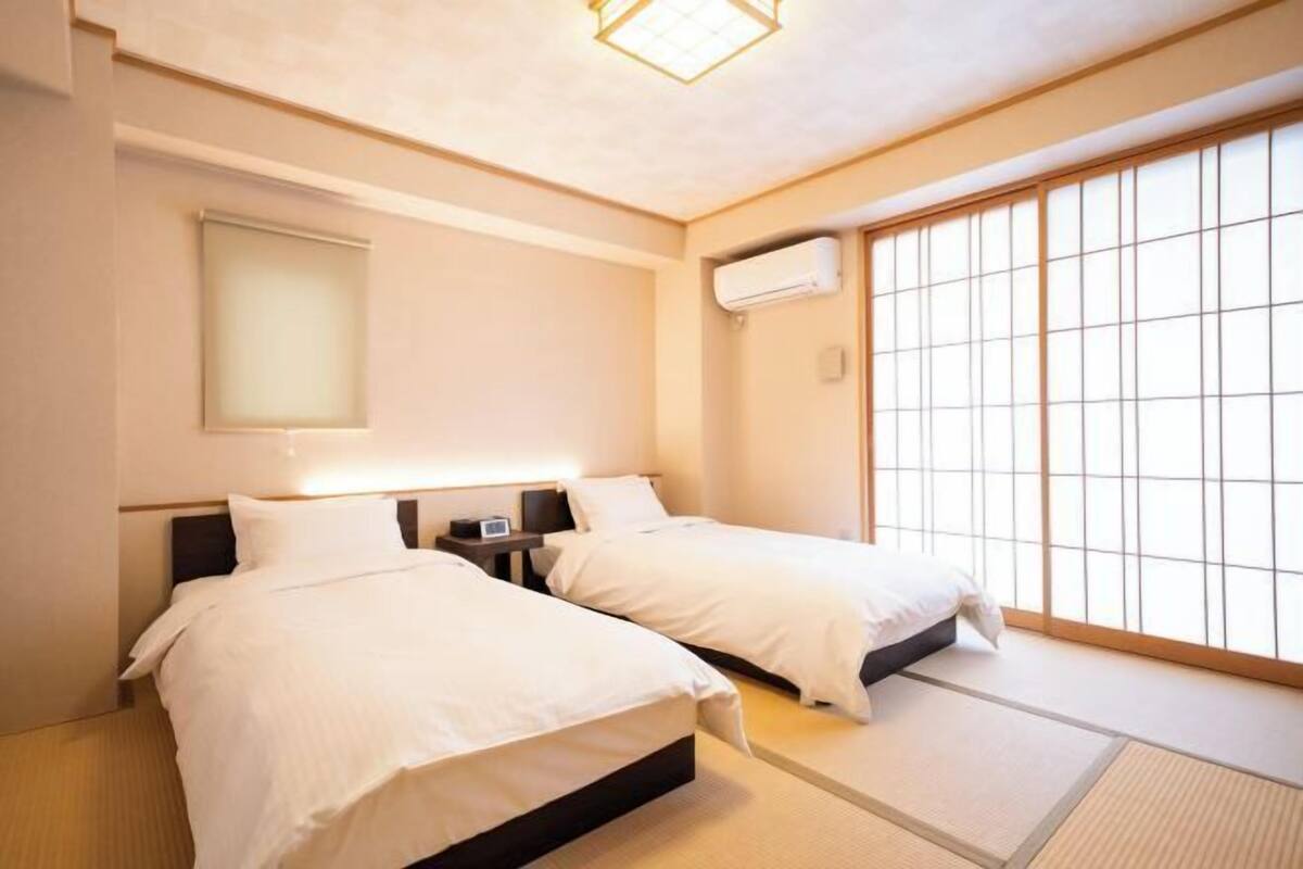 Habitación de hotel con dos camas, una mesita de noche, un aire acondicionado montado en la pared y una puerta corredera tipo shoji.
