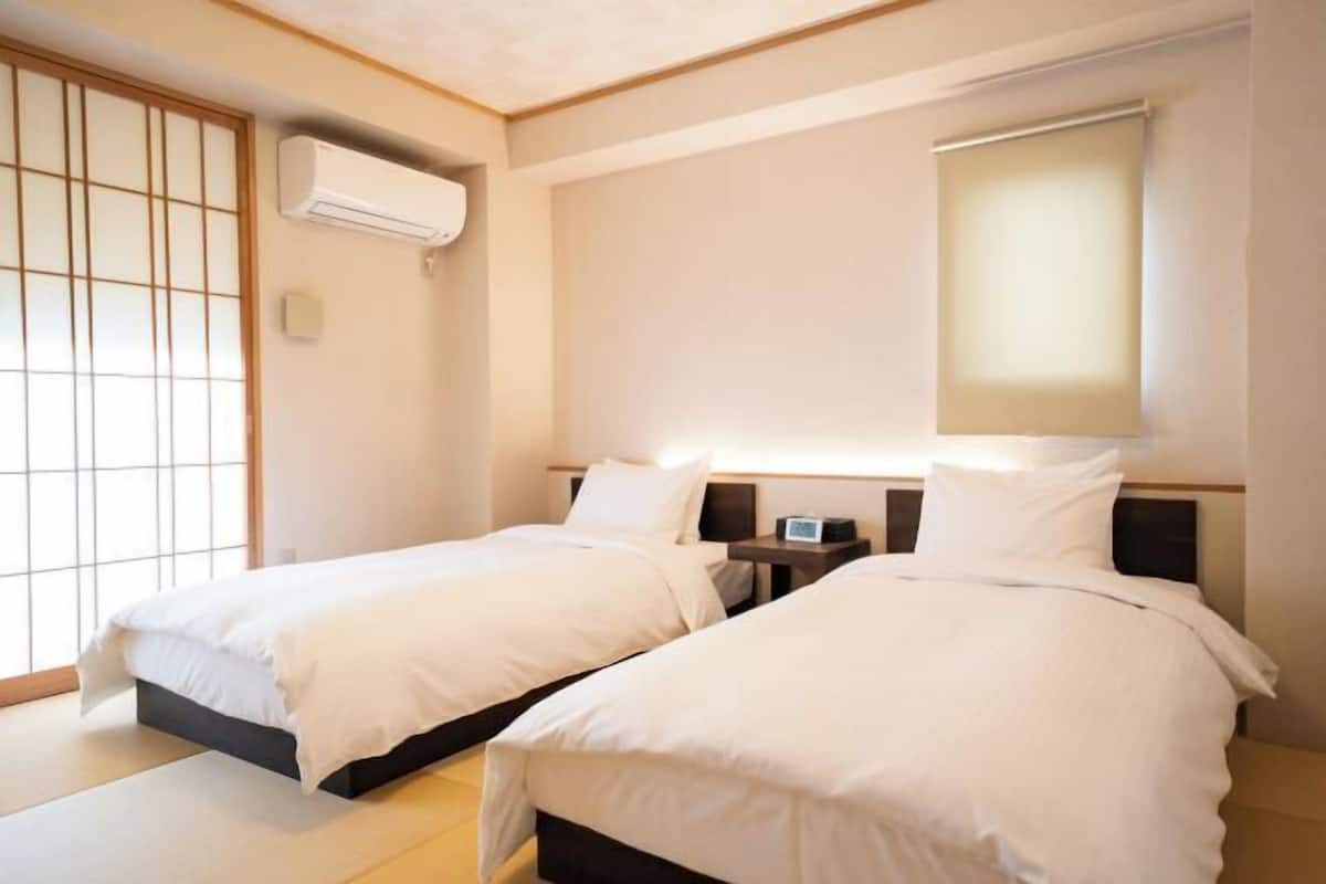 Habitación de hotel con dos camas, un panel corredizo tipo shoji, un equipo de aire acondicionado y una mesita de noche con lámpara.