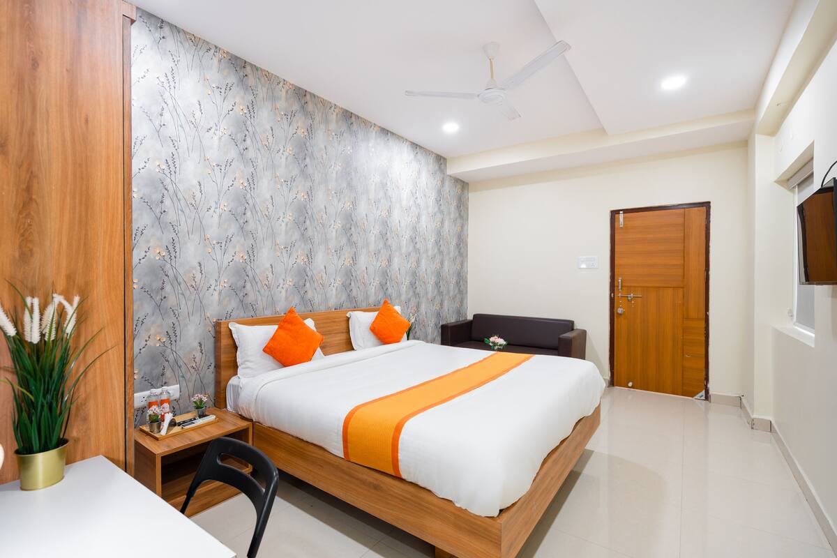 A modern hotel room with a wooden bed, orange pillows, a small table with a plant, and a TV on the wall.