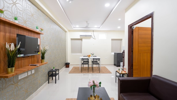 A modern living room with a flat-screen TV, a wooden entertainment unit, a dining area with a table and chairs, and a dark brown sofa. - Iris At Nest (Hyderabad)