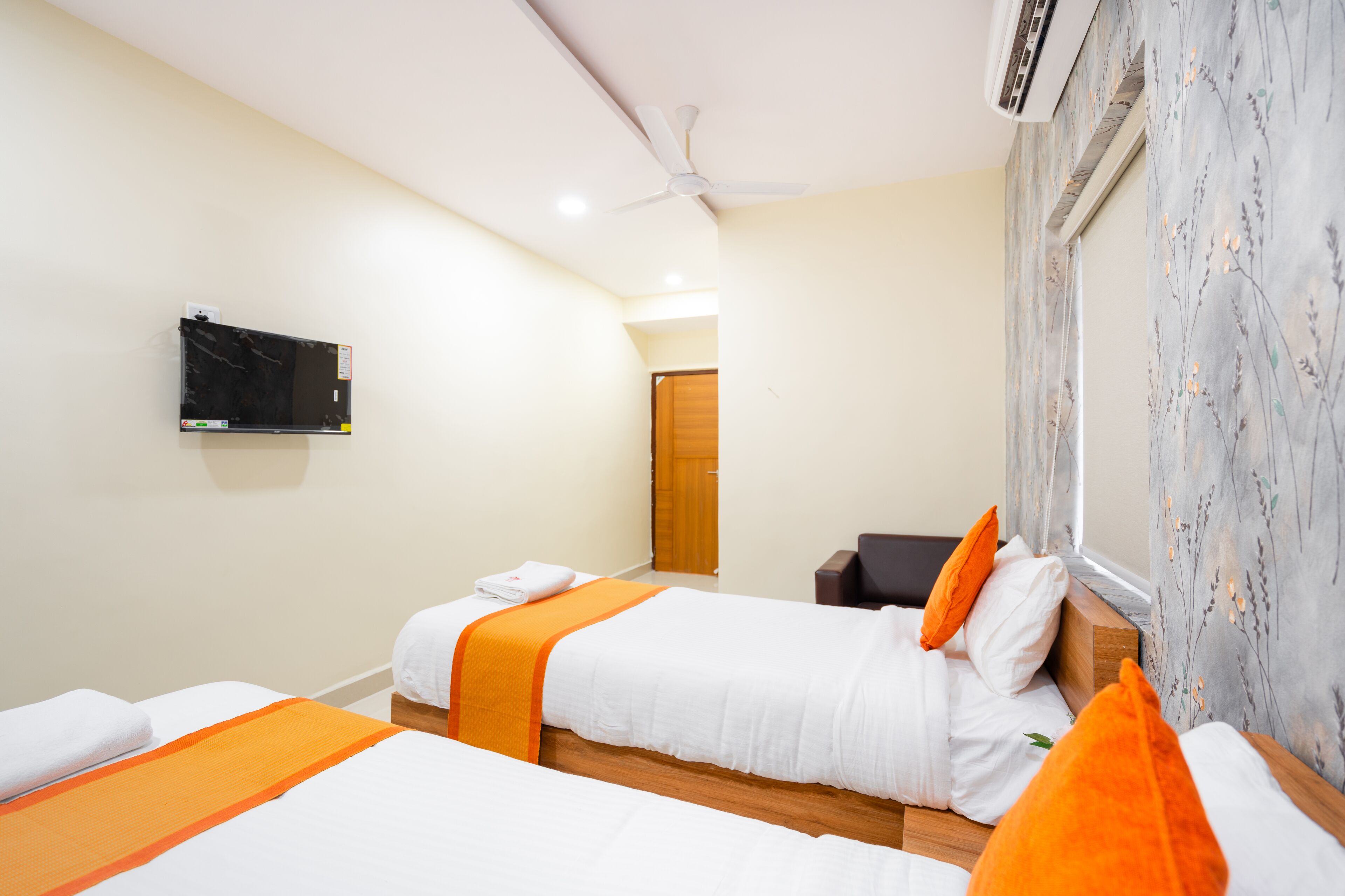 A hotel room with two beds, a flat-screen TV, a ceiling fan, and a window with floral curtains.