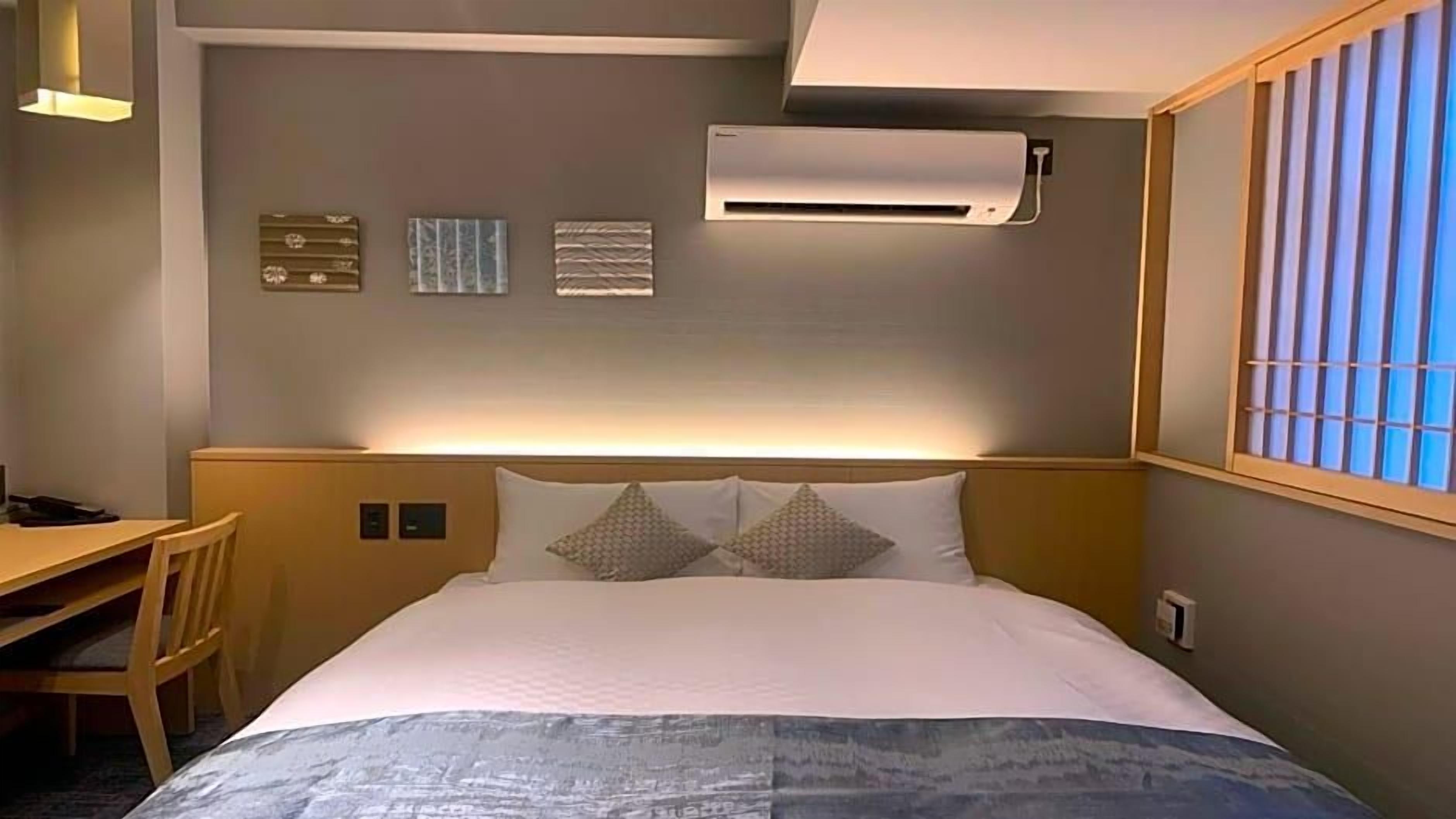 A hotel room with a bed, a desk, and a chair. There is an air conditioning unit on the wall and a window with blinds.