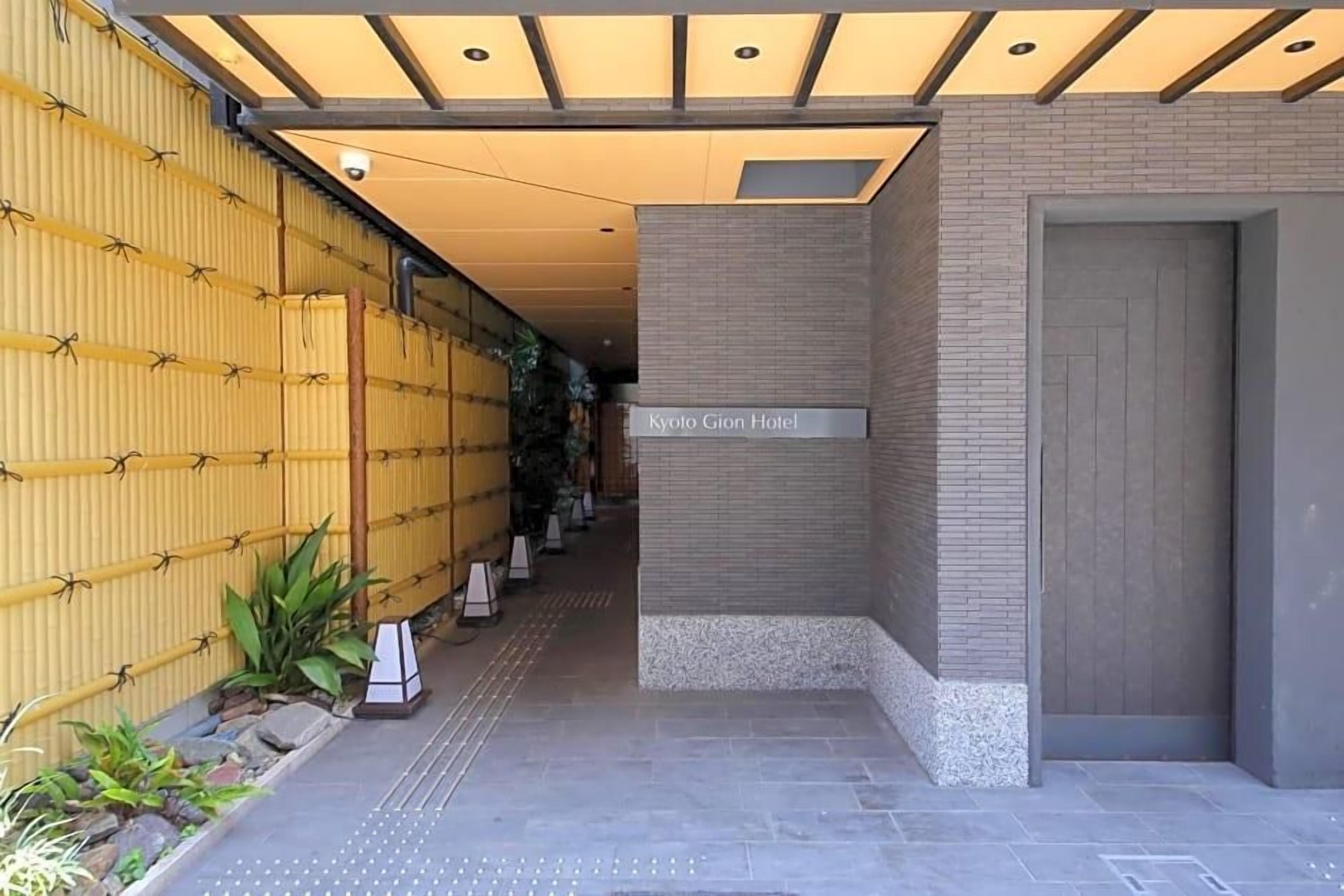 La entrada del Hotel Kyoto Gion, con un diseño moderno, presenta una pared de bambú y un camino despejado hacia la entrada.