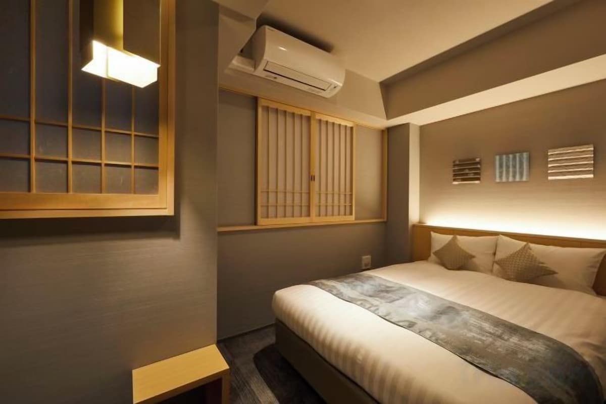 A hotel room with a bed, a window with wooden blinds, an air conditioning unit, and a bedside table.
