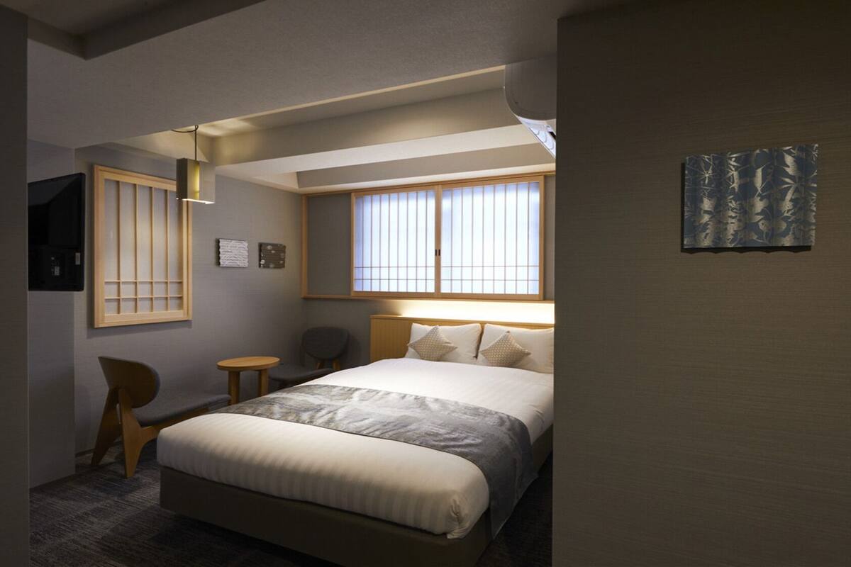 A hotel room with a bed, a chair, a small table, a window with blinds, and a wall-mounted artwork.