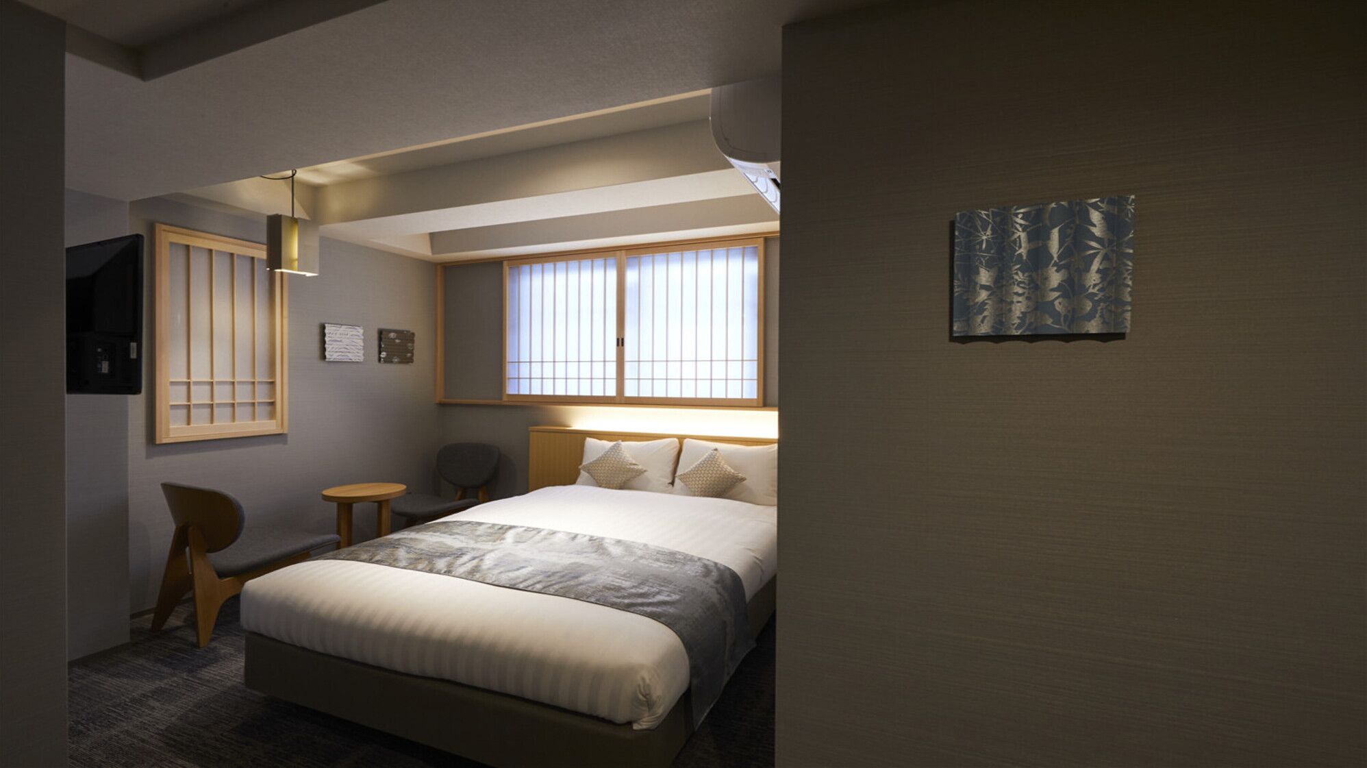 Une chambre d’hôtel comprenant un lit, une chaise, une petite table, une fenêtre avec des stores et une œuvre d’art accrochée au mur.