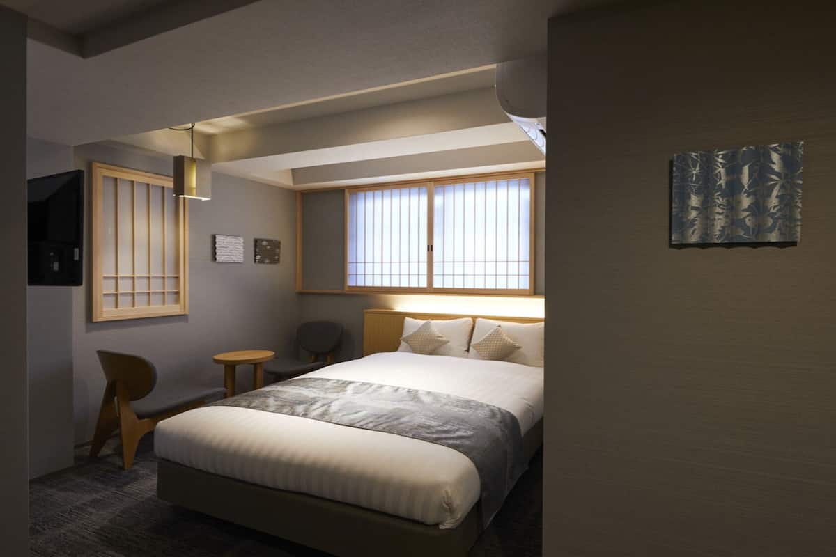 A hotel room with a bed, a chair, a small table, a window with blinds, and a wall-mounted artwork.
