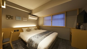 A hotel room with a bed, a desk, a chair, and a window with vertical blinds.