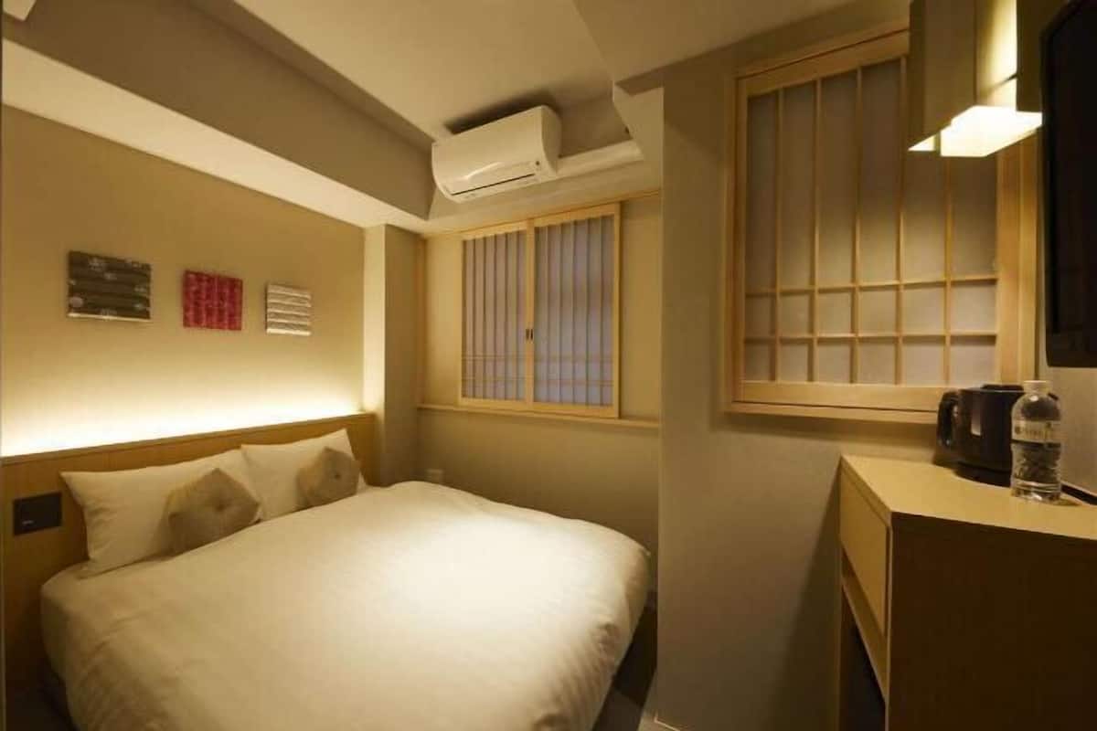 A hotel room with a bed, an air conditioner, a window with blinds, a bedside table with a bottle, and a wall-mounted clock.