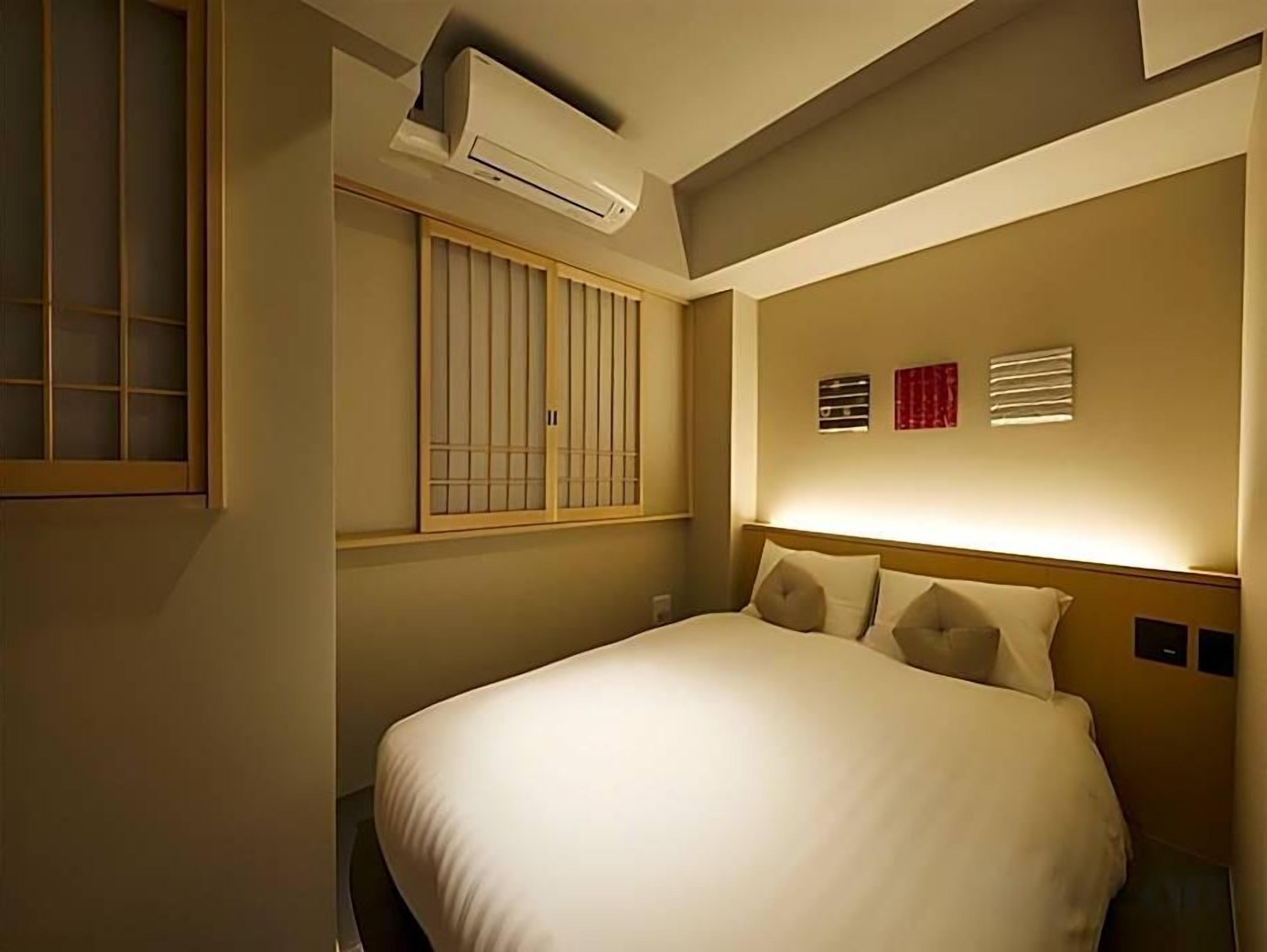 A hotel room with a bed, a window with blinds, an air conditioning unit, and a wall-mounted artwork.