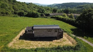 A rustic cabin with a metal roof, surrounded by lush greenery and hills in the background. - Charming Hut with Outdoor Bath & Decking, Sea Views (Sidmouth)