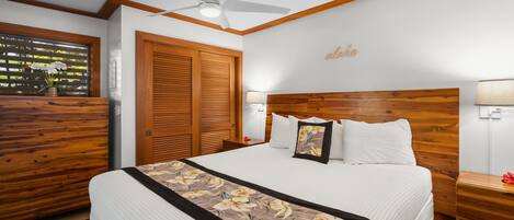 A bedroom with a wooden headboard, a bed with white bedding, a floral bedspread, a ceiling fan, and a window with a view of greenery.