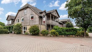 Een groot, twee verdiepingen tellend houten huis met een bakstenen fundering, omringd door een verharde oprit en weelderige begroeiing.