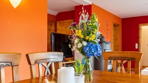 Een eettafel met een bloemenboeket en een keramieken haan, omringd door houten stoelen in een kamer met rode muren en een televisie op de achtergrond.