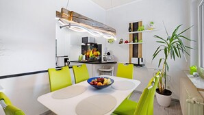 Un espace repas moderne avec une table blanche, des chaises vertes, un saladier de fruits et une plante en pot.
