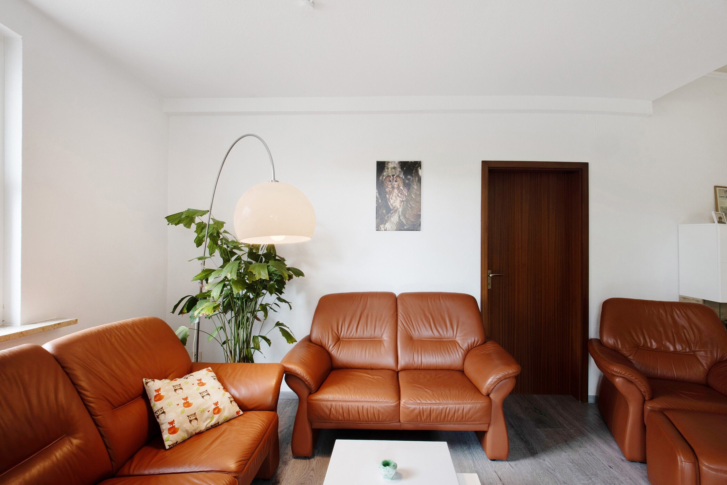 Un salon avec un canapé en cuir marron, une table basse blanche, un lampadaire et une porte en bois.