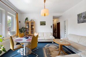 Ett vardagsrum med en vit soffa, ett matbord med gula stolar, ett soffbord, en hängande lampa och en bokhylla.