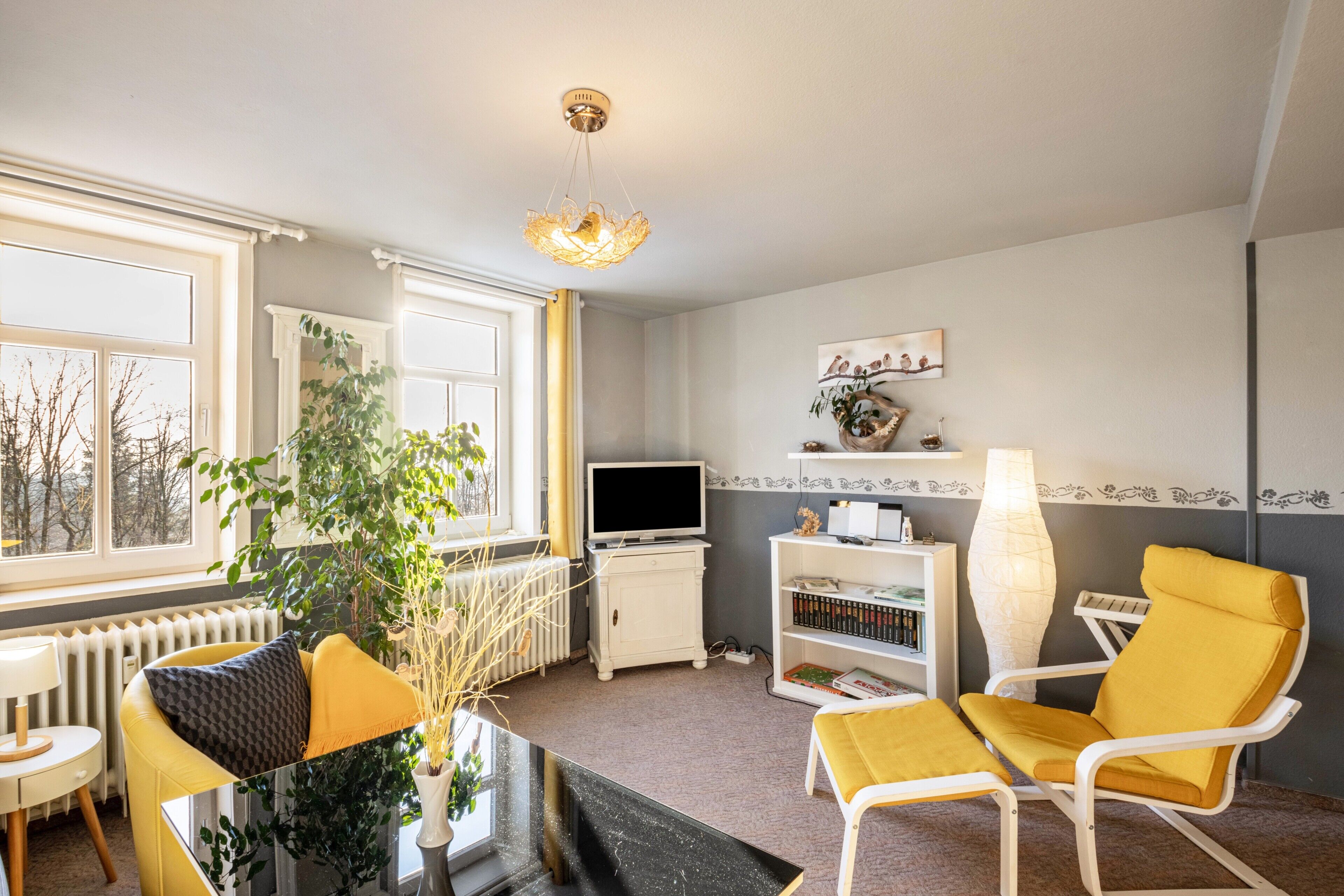 Ein Wohnzimmer mit Fernseher, Bücherregal, gelben Sesseln, einer Pflanze und einem Fenster.