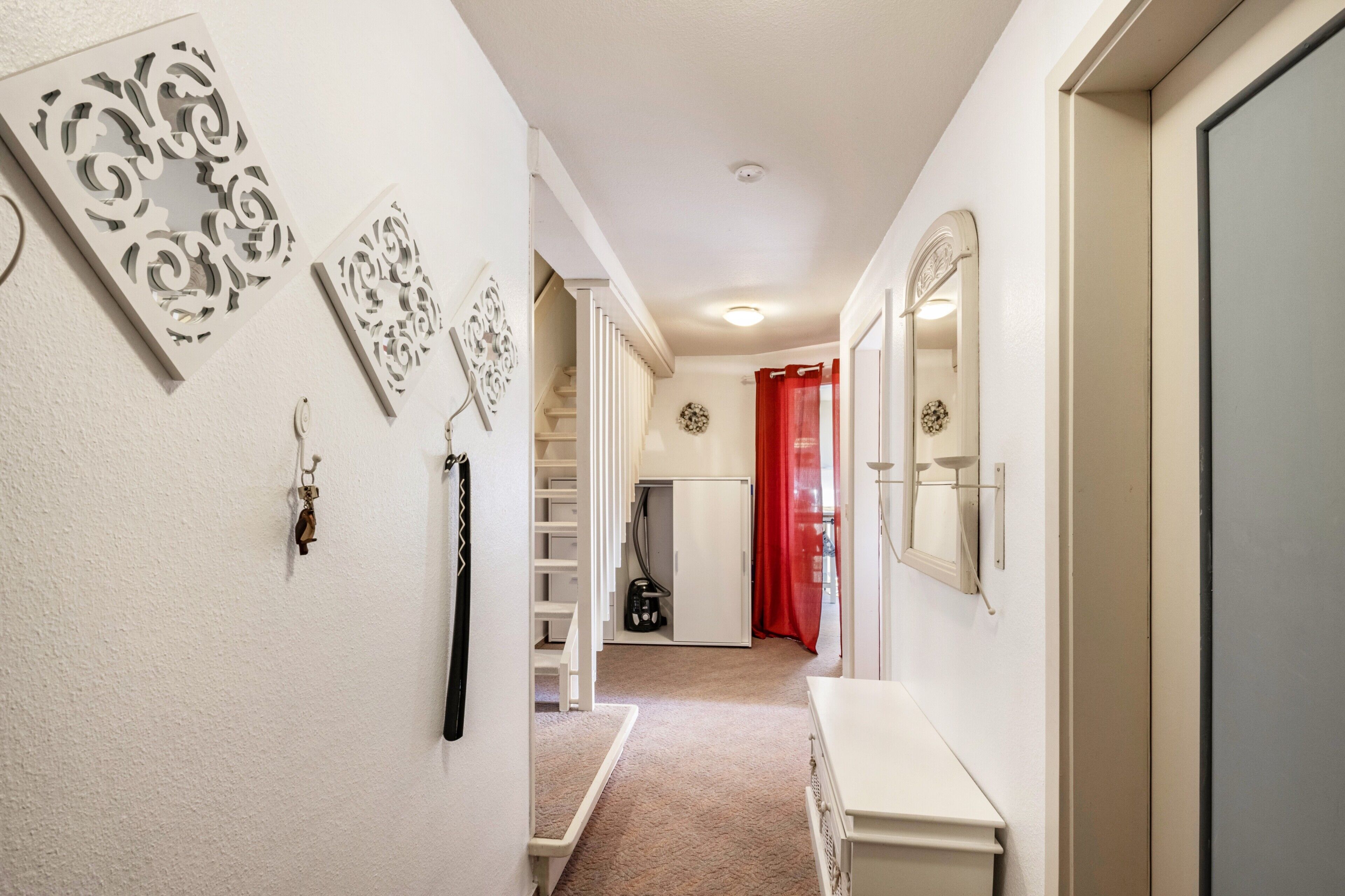 Un pasillo con pared blanca, un banco blanco, una cortina roja y una puerta blanca.