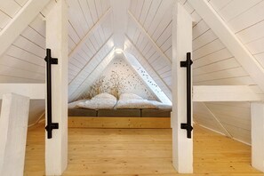 Une chambre à coucher aménagée dans un espace mansardé, avec un plafond en pente, des poutres en bois et un lit agrémenté d’oreillers à motifs.