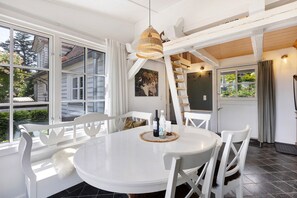 Un espace de salle à manger aux tons clairs, avec une table ronde, des chaises blanches, un luminaire suspendu et une vue sur l’extérieur grâce à de grandes fenêtres.