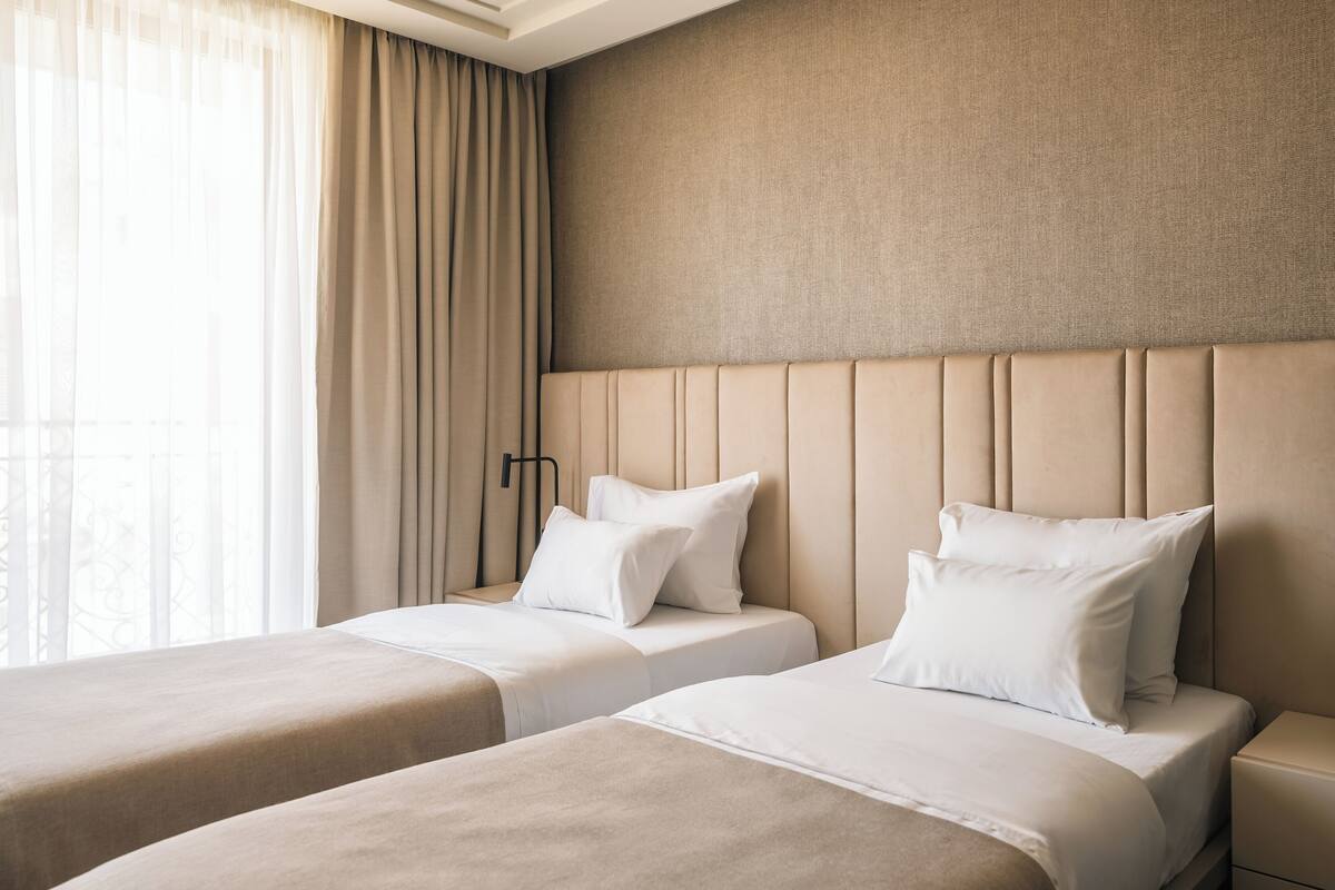 A hotel room with two beds, beige bedding, a large window with curtains, and a plain wall.