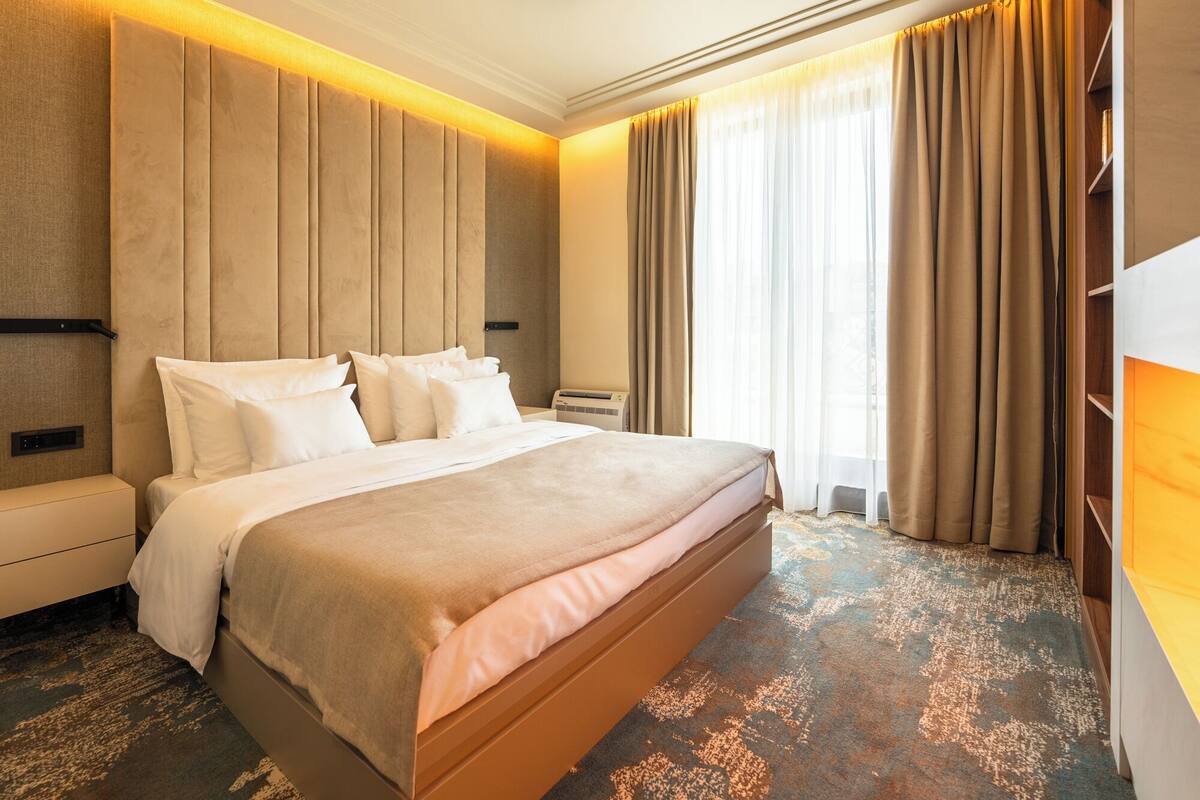 A hotel room with a large bed, a headboard with vertical panels, a bedside table, a nightstand, a built-in air conditioning unit, and a large window with curtains.