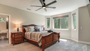 Ein Schlafzimmer mit einem hölzernen Bett, einem Deckenventilator, einem Nachttisch, einer Lampe und großen Fenstern.