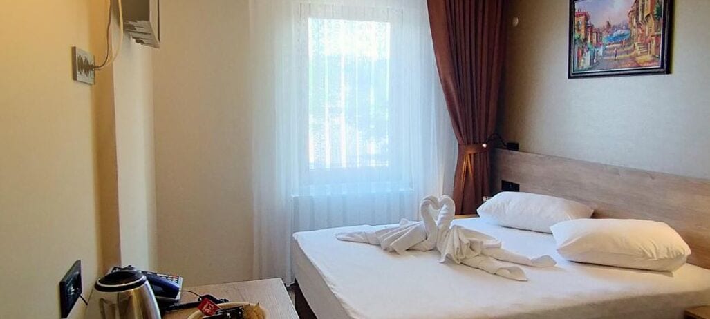 Una habitación de hotel con cama, una ventana con cortinas traslúcidas, un cuadro en la pared y un perchero.