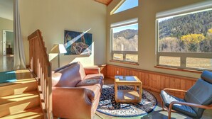 A cozy living room with a brown leather sofa, a wooden coffee table, and a view of a forested hillside through large windows. - Close to Hot Springs: Chalk Creek Meadows Home! (Nathrop)