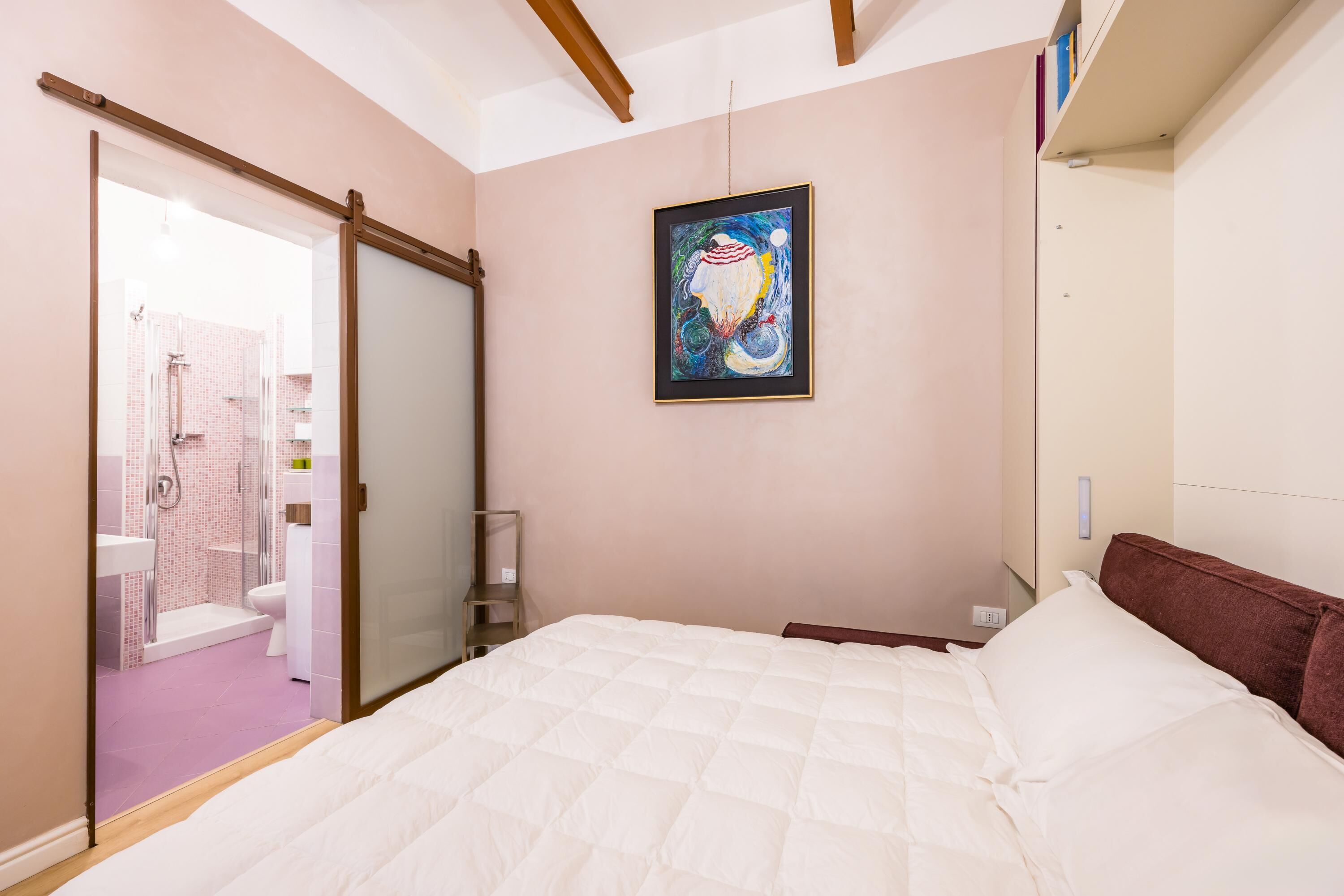 Un dormitorio con cama, un cuadro en la pared y un baño visible a través de una puerta corredera.