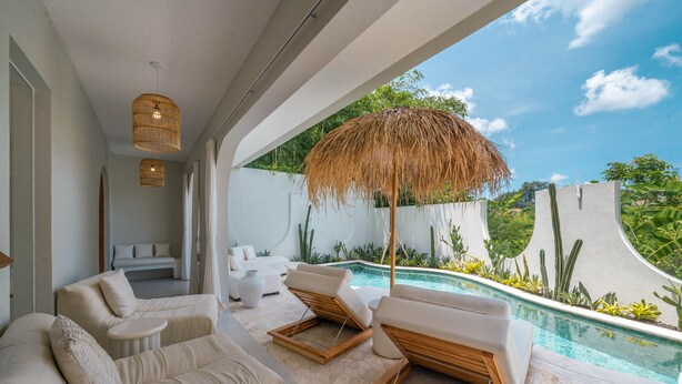 A modern outdoor lounge area with a pool, white sofas, and a thatched umbrella.