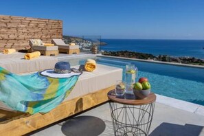 A poolside area with a lounge chair, a small round table with fruits and a bottle, and a view of the sea. - Sunny Drops Villa White by Estia (Mylopotamos)