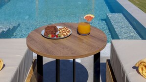 A poolside table with a plate of food and a drink, overlooking a pool and a wooden pergola. - Sunny Drops Blue Villa by Estia (Mylopotamos)