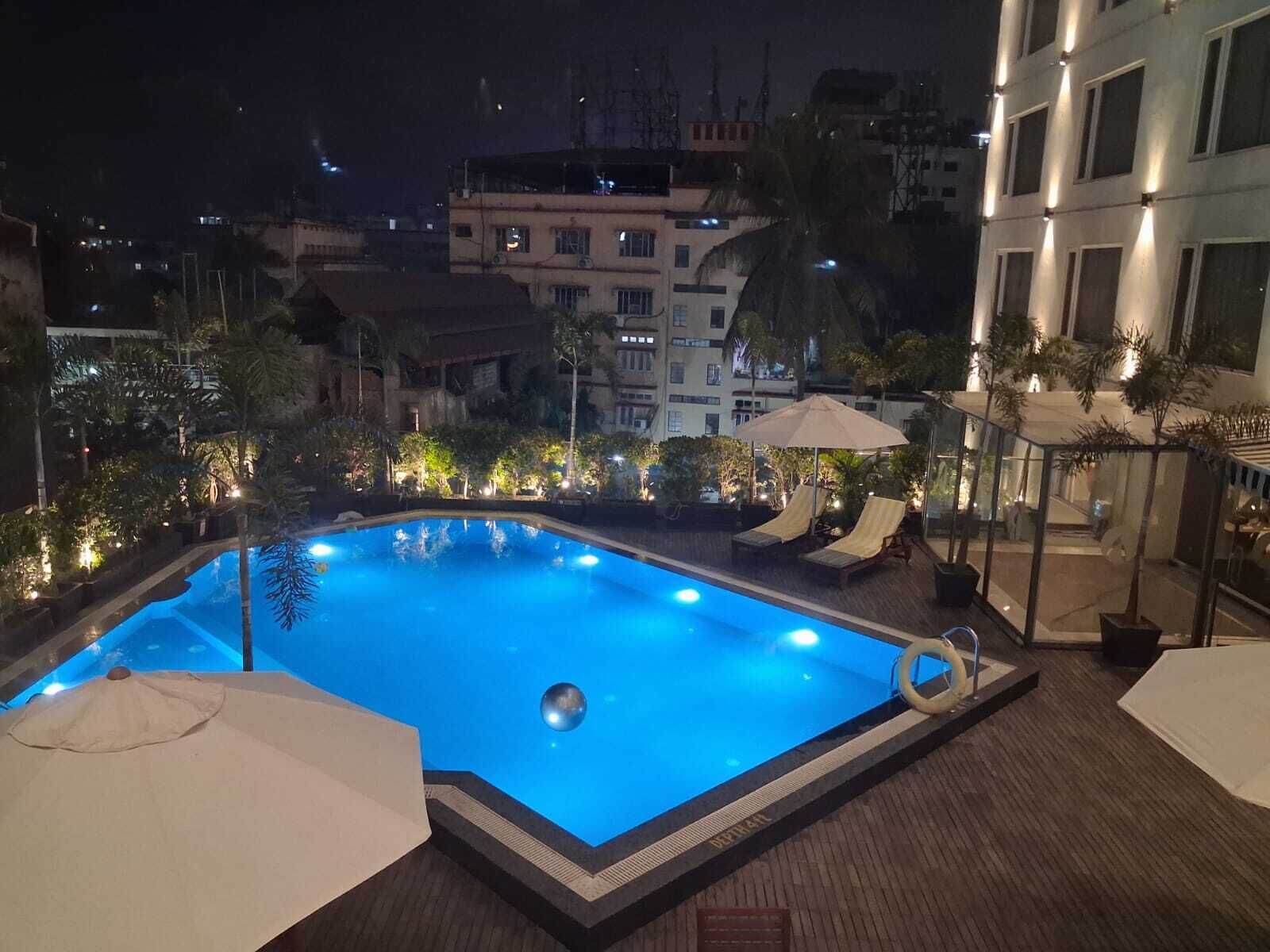 A rooftop swimming pool with lounge chairs and umbrellas, surrounded by buildings and trees.