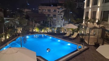 A rooftop swimming pool with lounge chairs and umbrellas, surrounded by buildings and trees.