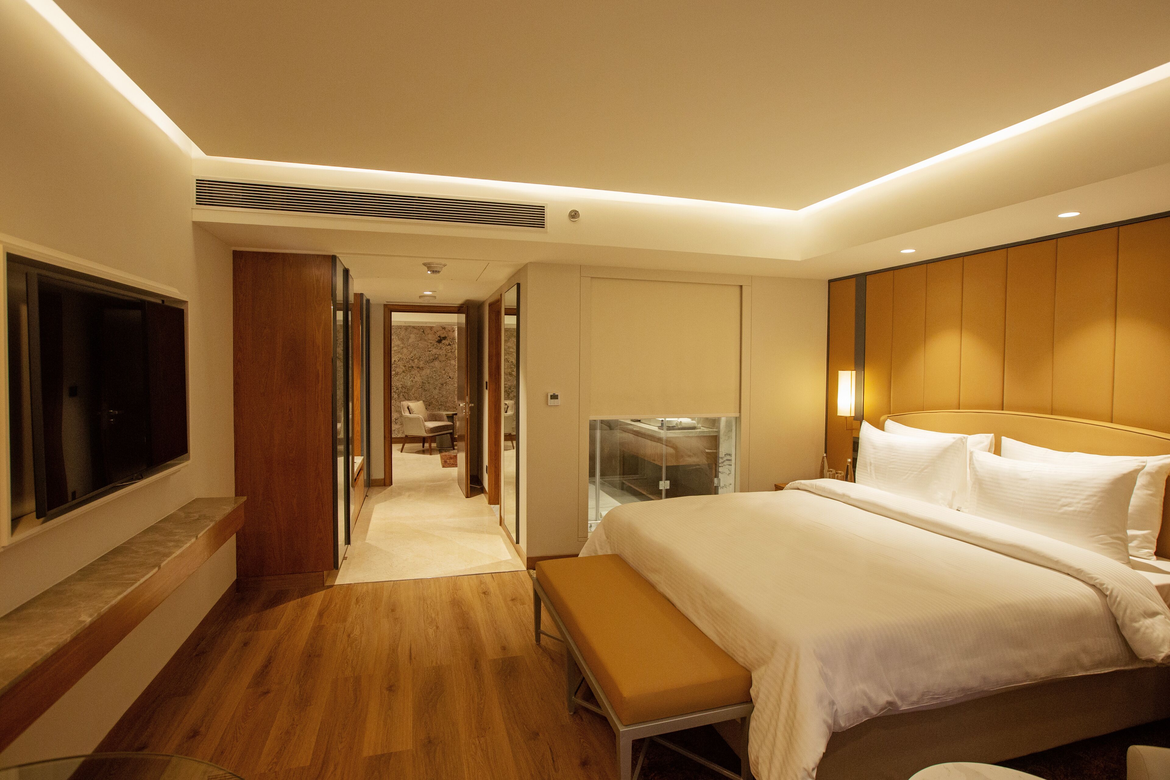 A modern hotel room with a large bed, a bench, a TV, and a walk-in closet.