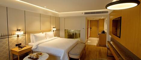 Una habitación de hotel moderna con una cama grande, mesitas de noche, una pequeña mesa redonda con una bandeja de aperitivos, un banco, una mesa de centro de cristal y un televisor montado en la pared.