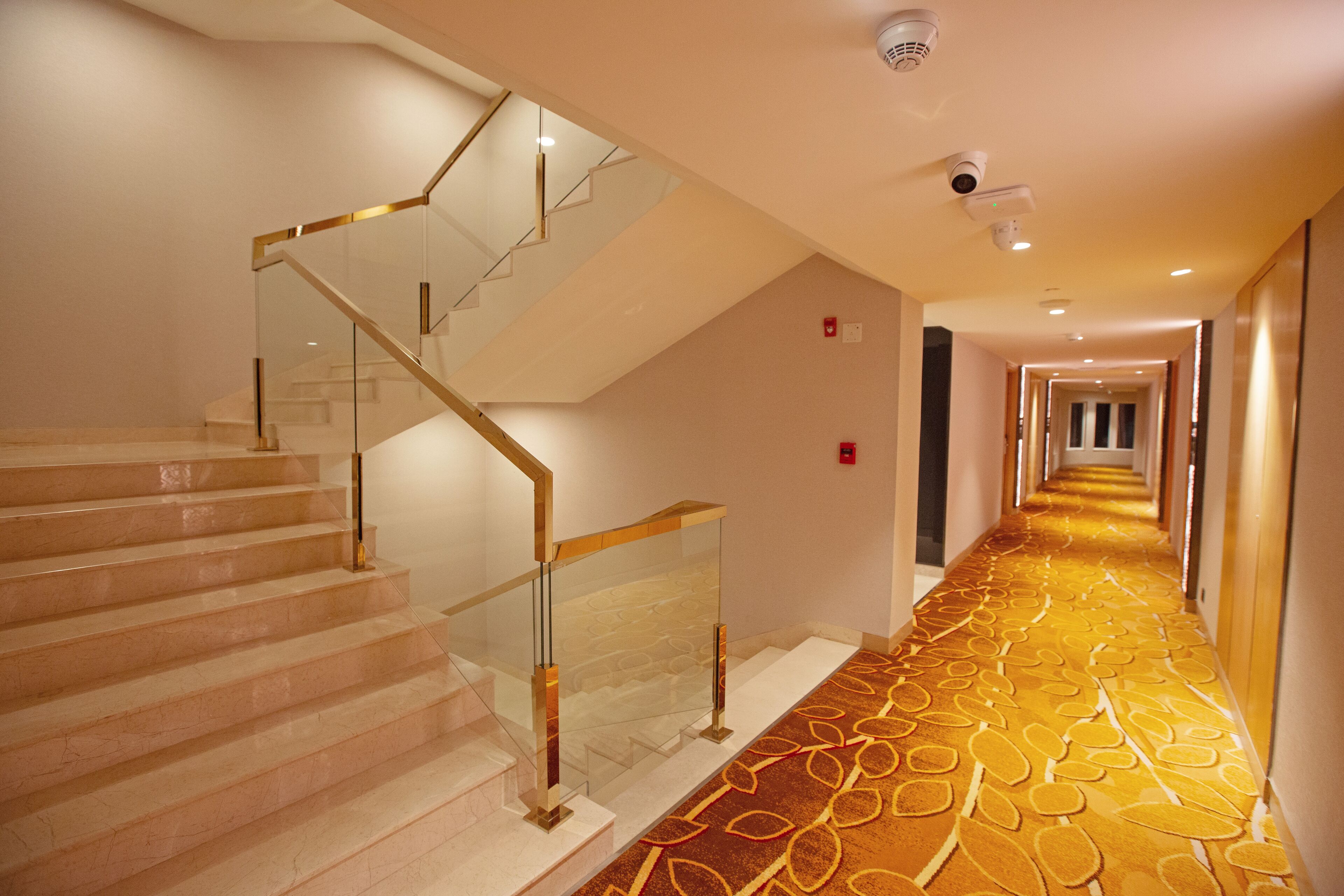 A hotel hallway with a carpeted floor, a glass railing staircase, and a security camera on the ceiling.