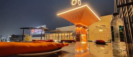 A rooftop dining area with a large illuminated sign, outdoor seating, and a view of the cityscape.