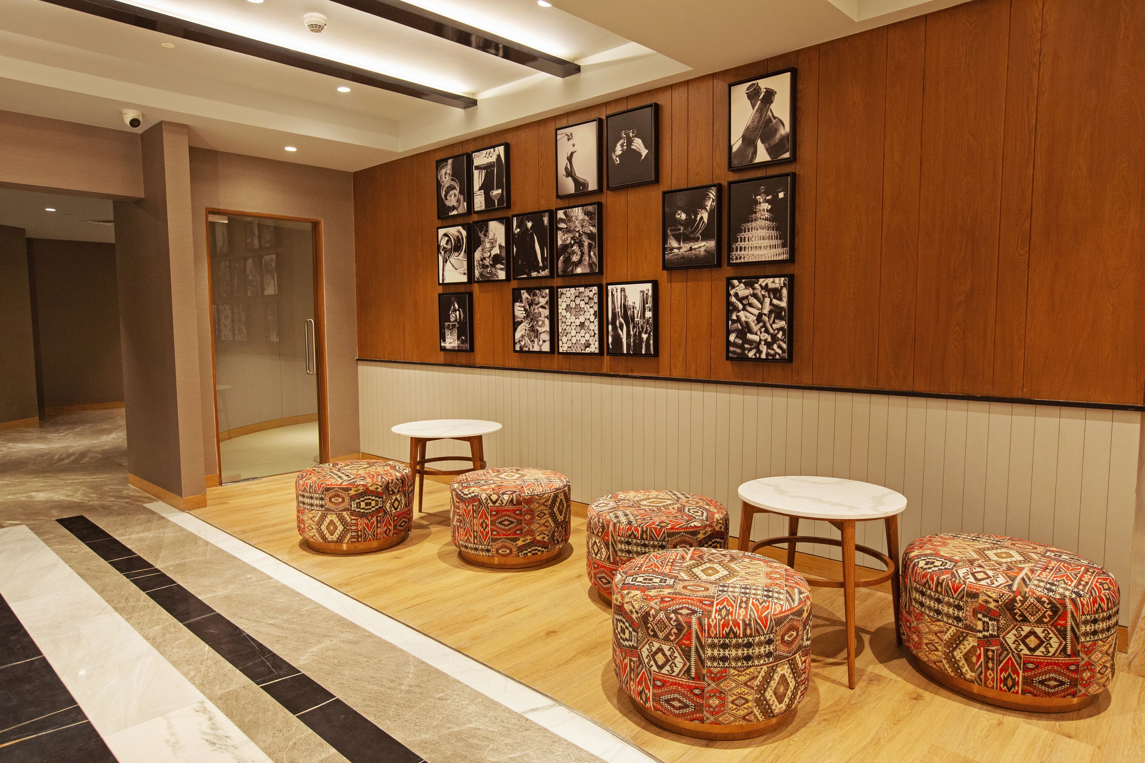 Un hall d’entrée avec des murs lambrissés en bois, une porte en verre et un sol carrelé. On y trouve quatre poufs à motifs et trois tables blanches.