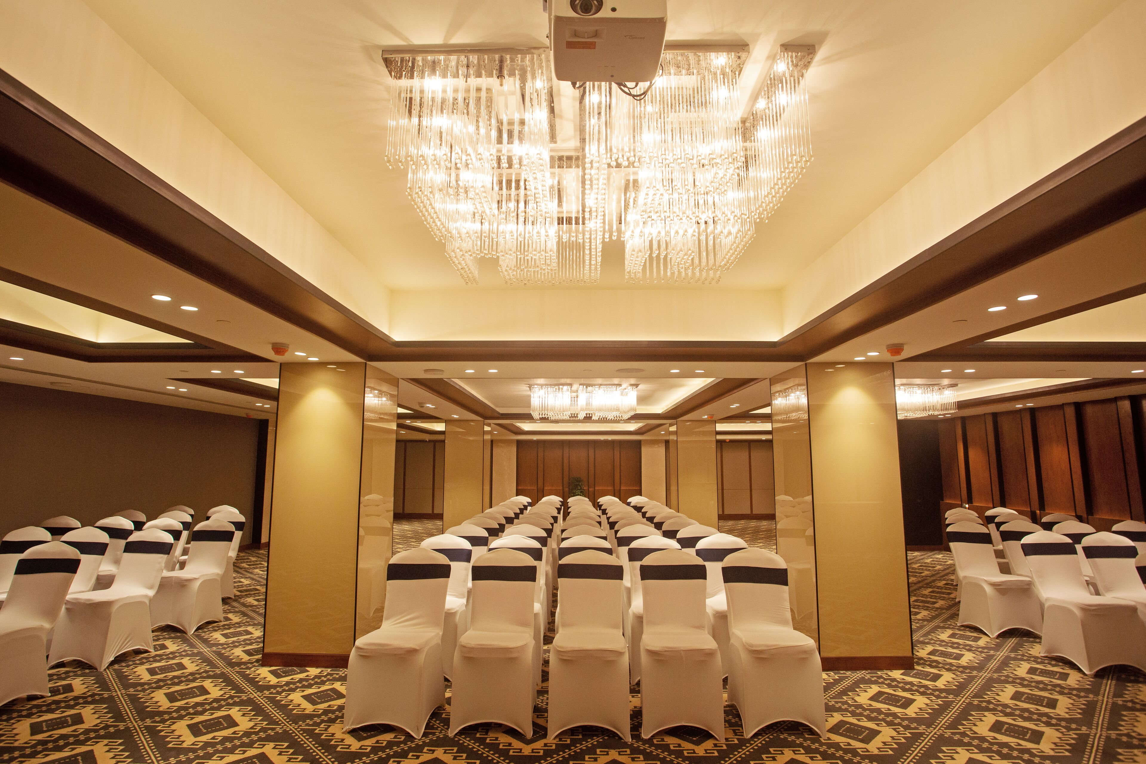 A conference room with rows of white chairs, a large chandelier, and patterned carpet.