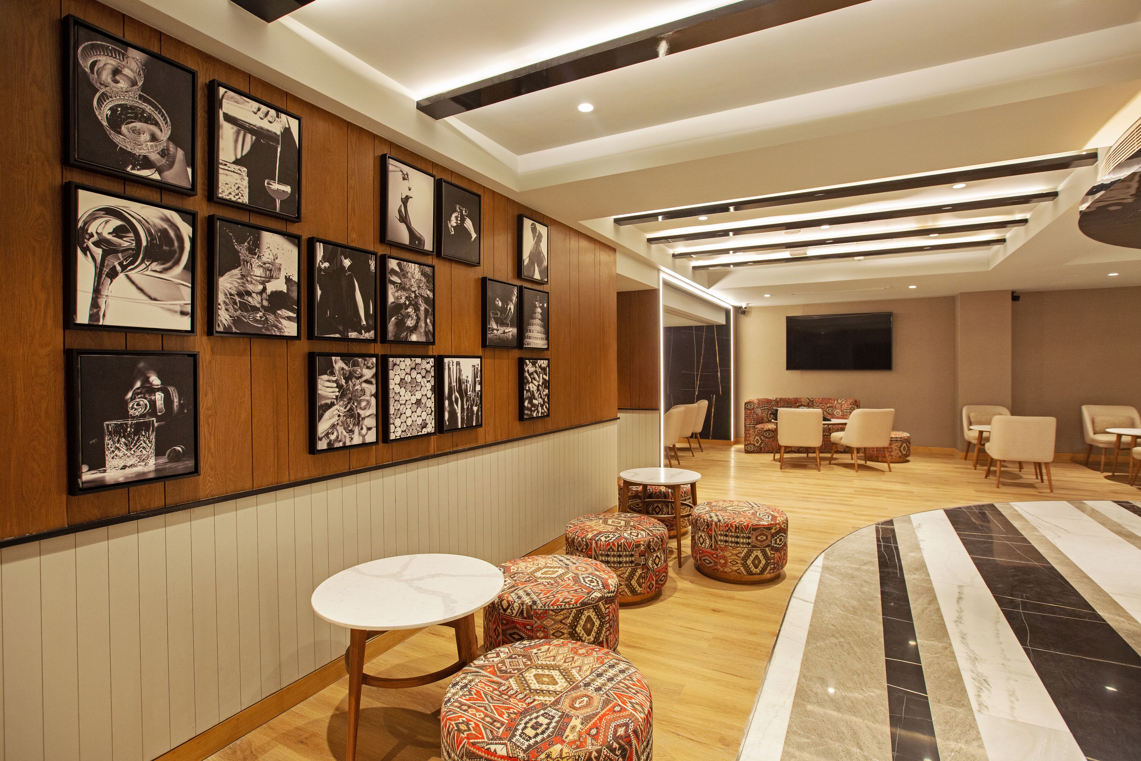 A modern hotel lobby with a wooden panel wall adorned with black and white photographs, a seating area with round tables and patterned ottomans, and a large curved marble table.