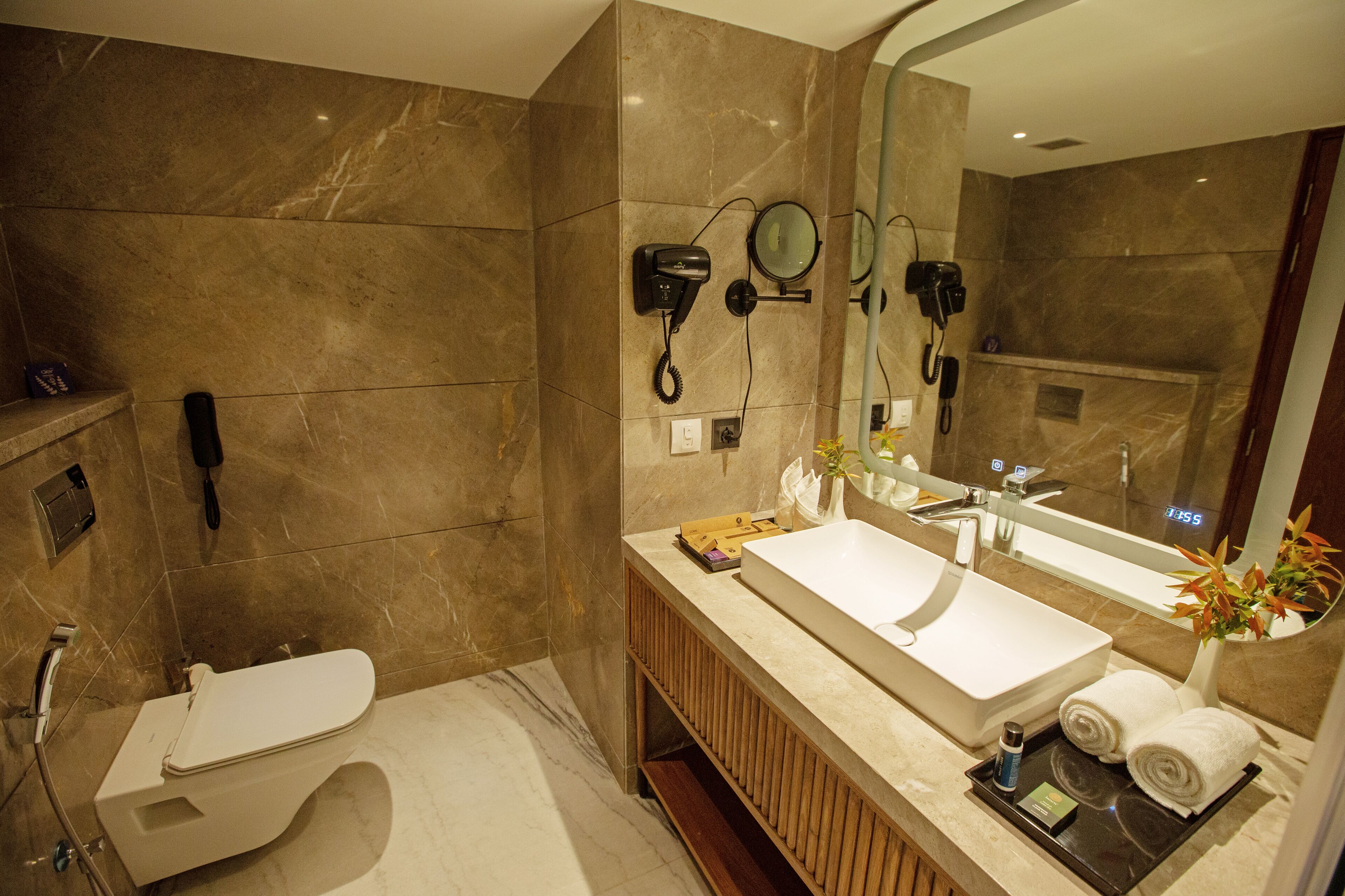 A modern bathroom with a large mirror, a sink, and a toilet. There are hair dryers mounted on the wall, and a phone is also visible.