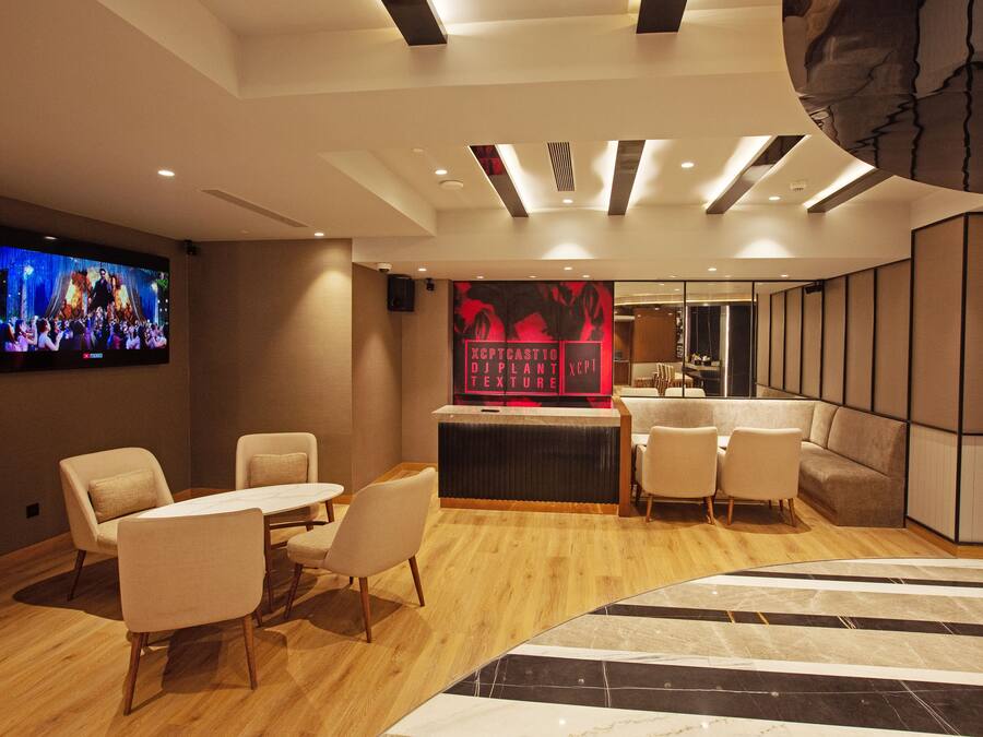 Un vestíbul d'hotel modern amb taulell de recepció, zona de seients i un gran televisor de pantalla plana que mostra un concert.